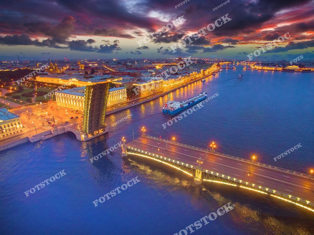На фотографии изображен Троицкий мост, разведенный над Невой для прохода судов. Вечерняя подсветка подчеркивает его архитектуру и делает пейзаж особенно эффектным. На заднем плане виднеются здания исторического центра Петербурга, включая силуэт Исаакиевского собора и другие памятники города. Окруженный огнями мост и река отражают романтику ночного Санкт-Петербурга.
